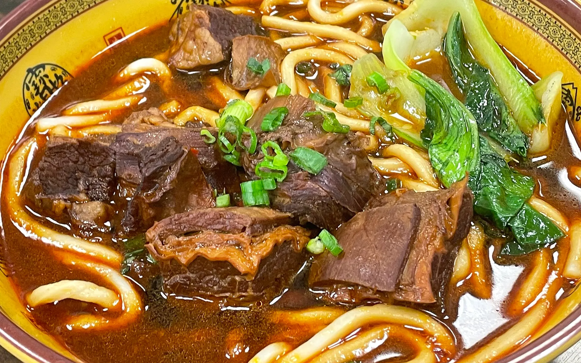 A bowl of braised beef noodles — wide noodles, brisket and greens in a yellow fretwork bowl