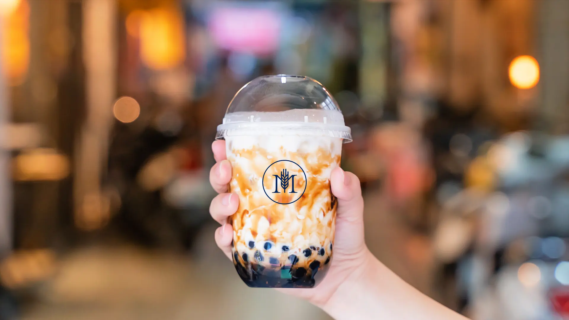 A hand holds a brown-sugar bubble tea, the M Chá Bar line mark on the cup, against the blurred lights of an evening street