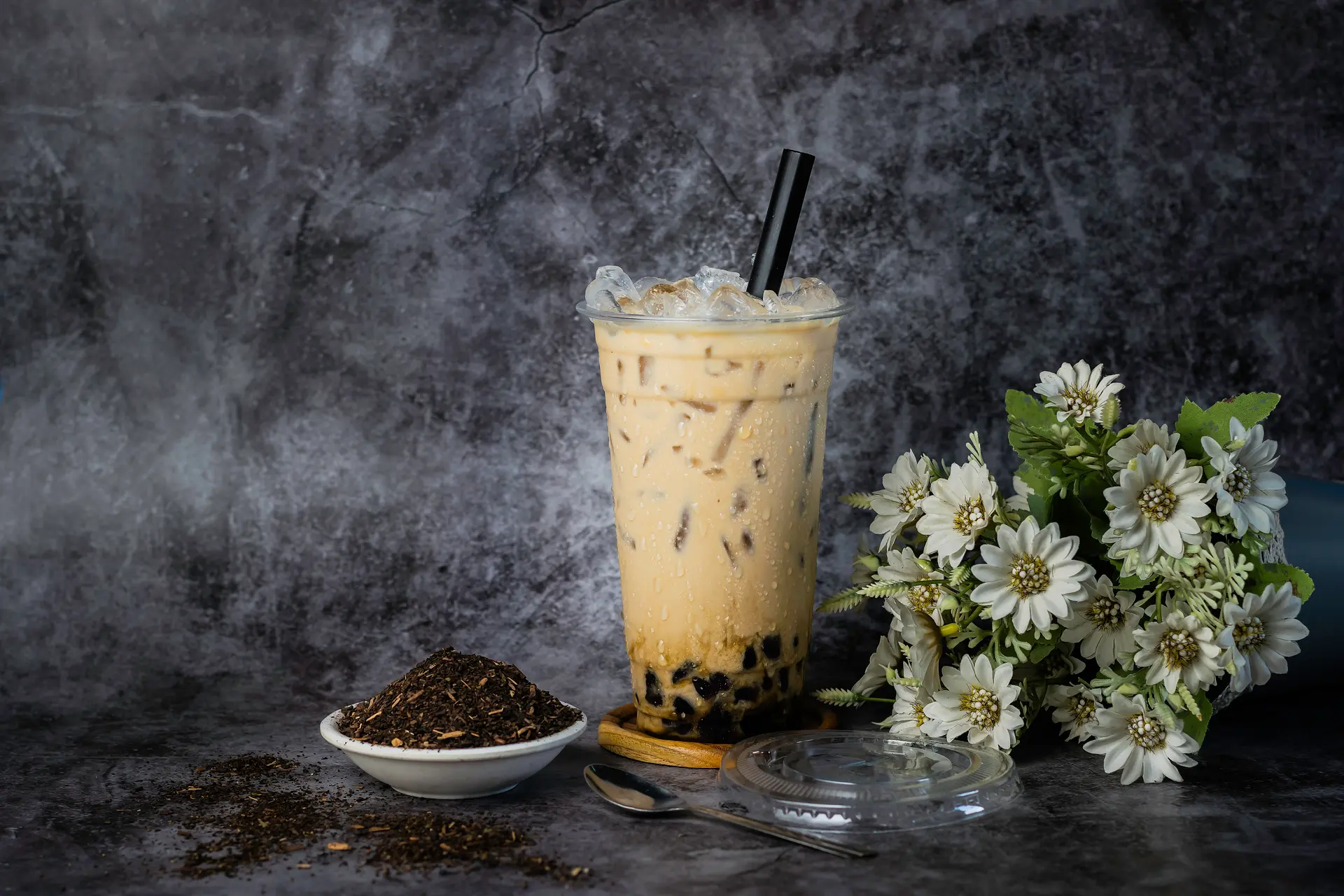 A milk tea, a dish of tea leaves and a spray of white daisies on dark stone