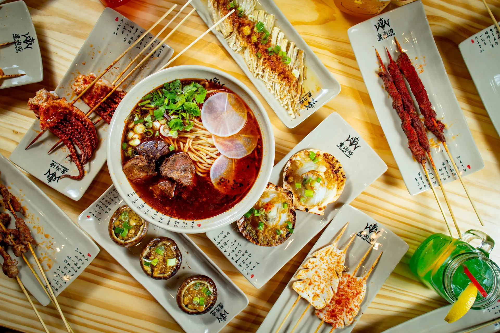 From above: a bowl of beef noodles at the centre of a table of skewers, mushrooms, small plates and fruit drinks, on the restaurant’s own plates
