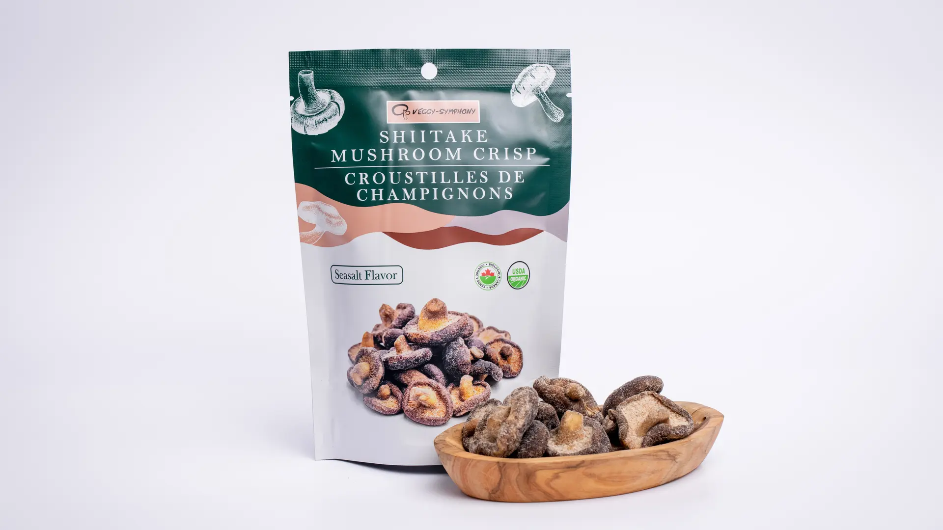 The printed shiitake crisp pouch beside a wooden bowl of crisps on pale grey